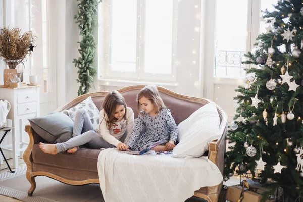 Deux filles assises sur le canapé et feuilletant et lisant un livre, deux sœurs, la chambre est décorée pour la nouvelle année et Noël — Photo