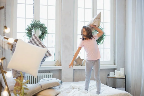 Deux filles, sœurs combattant des oreillers sur le lit, la fenêtre décorée d'une couronne de Noël, la vie, l'enfance — Photo