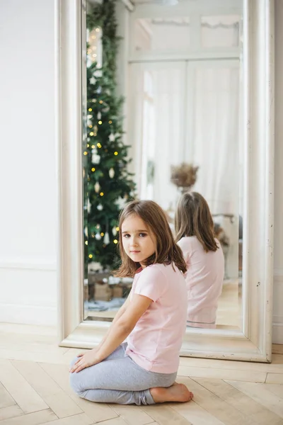 La fille est assise près du miroir, le miroir reflète l'arbre de Noël et les lumières, l'intérieur est décoré pour la nouvelle année, Noël, attendant les vacances — Photo