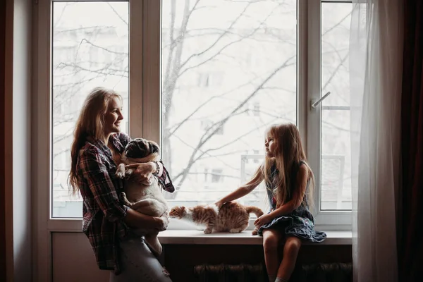 Mom holds a dog and stands near the window, the girl sits near the window, the family home