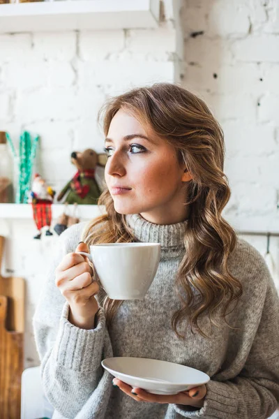 Avrupa görünümlü bir kız mutfakta oturuyor, beyaz bir fincandan çay ya da kahve içiyor, içiyor, sabah mutfakta, öğrenci, müdür, serbest yazar. — Stok fotoğraf