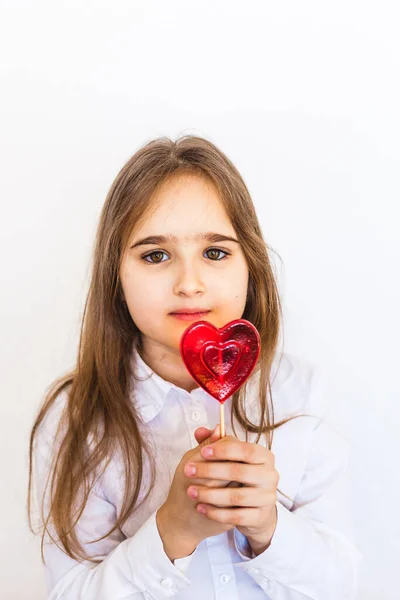 Uma menina de aparência europeia em um fundo branco tem um pirulito em forma de coração em sua mão, amor, presente, família, Dia dos Namorados — Fotografia de Stock
