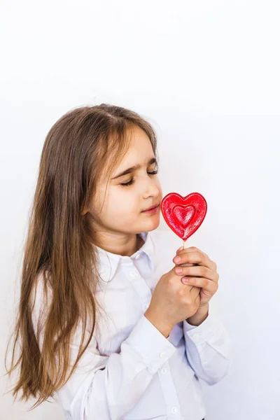 Una chica de aspecto europeo sobre un fondo blanco sostiene una piruleta en forma de corazón en su mano, amor, regalo, familia, día de San Valentín —  Fotos de Stock
