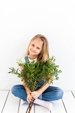 Avrupalı görünümlü uzun saçlı bir kız yerde oturuyor ve elinde şam fıstığı dalları, yeşil dallar, bitkiler, iç kısımlar var.
