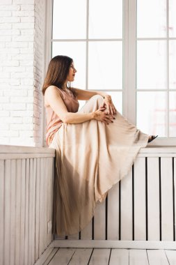 Açık renk elbiseli bir kız, pencerenin kenarında oturuyor, geleceği düşünüyor, evi, sokakları, yürüyüşleri, yalnızlığı, kendini önemsemeyi, gelenekleri ve kınayı.