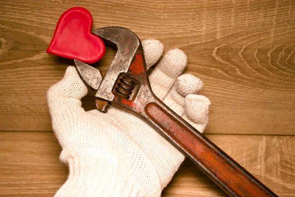 Valentine\'s Day. Red heart in male hands in gloves held by an adjustable wrench. On a wooden background. Concept for design.