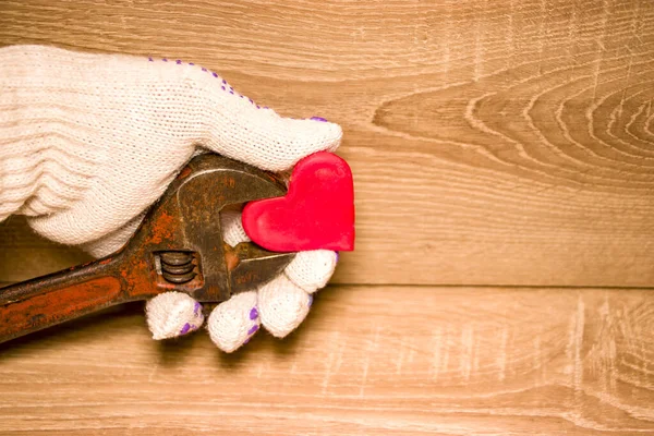 Valentine\'s Day. Red heart in male hands in gloves held by an adjustable wrench. On a wooden background. Concept for design.