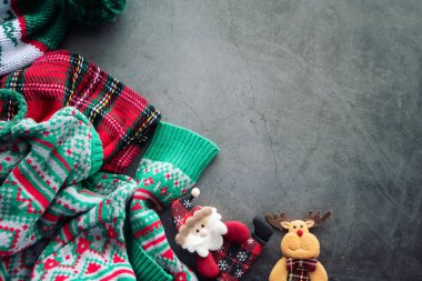 Yeşil kazak ve kırmızı İskoç battaniyesi çimento arka planda Noel Baba bebeği ve fotokopi alanı olan bir ren geyiği ile duruyor.