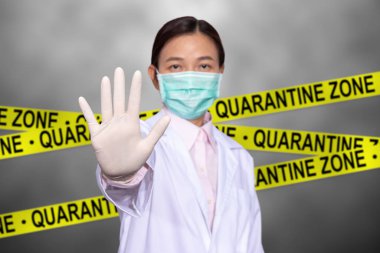 Asyalı kadın doktor tıbbi maske takın, dur işareti için elinizi kaldırın. Arkasında sarı karantina işareti olan karantina bölgesine girmeyin. Karantina bölgesinde giriş yasaktır..