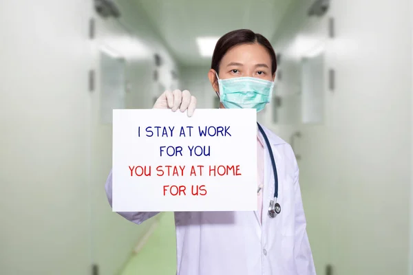 医院里的亚洲医生戴着口罩 拿着写着 我在为你工作 你呆在家里为我们工作 的文件 呆在家里开展政策运动 以控制Covid 19禽流感疫情的爆发 — 图库照片