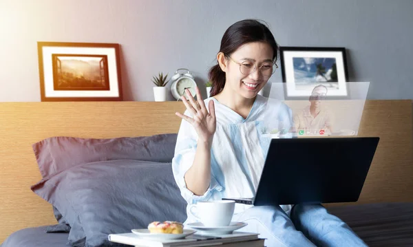 attractive happy woman using video call, make online conversation via laptop computer on bed from her room, having video chat, waving hand with visual screen of conversation partner in pop-up window