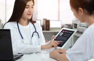 Tıbbi muayene odasındaki hastaya danışılırken ekranda hasta tıbbi kaydı olan Asyalı kadın doktor elinde tablet tutuyor..