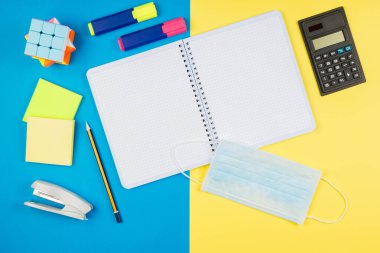 Karantinadan sonra okula dönmek, tecrit. Okul malzemeleri, tıbbi maske ve sarı ve mavi arka planda eldivenler. Sarı üzerine izole hesap makinesi, makas, kalem, defter.