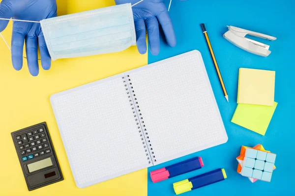 Back to school after quarantine, isolation. School supplies, medical mask and gloves on yellow and blue background. Calculator, scissors, pen, notebook isolated on yellow.