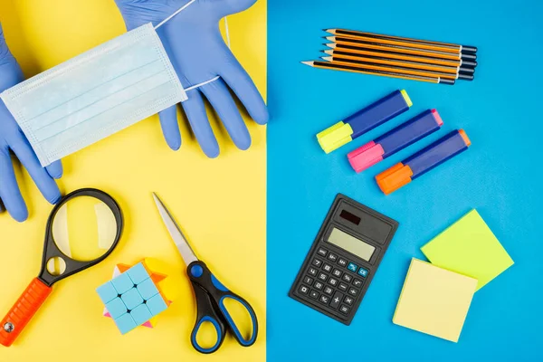 Back to school after quarantine, isolation. School supplies, medical mask and gloves on yellow and blue background. Calculator, scissors, pen, notebook isolated on yellow.