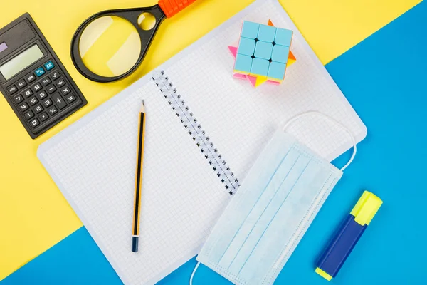 Back to school after quarantine, isolation. School supplies, medical mask and gloves on yellow and blue background. Calculator, scissors, pen, notebook isolated on yellow.