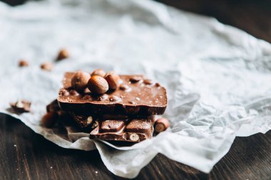 Fındıklı sütlü çikolata: Fıstık, beyaz parşömen kağıdında fındıklı fındık ve siyah ahşap masa. Seçici odak