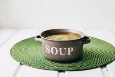 Tavuk ve sebzeli lezzetli sebze çorbası: brokoli, havuç, soğan ve baharat. Sağlıklı yiyecekler. Çorba tabağı. Demir kaşık tabakta yatıyor. Bir tabak yeşil halıdaki pencerenin yanında duruyor.