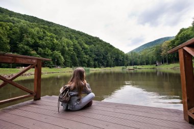 Kız, tahtadan bir iskelenin kenarında oturmuş gölün kenarında dinleniyor. Güneşli yaz günü veya akşam konsepti.