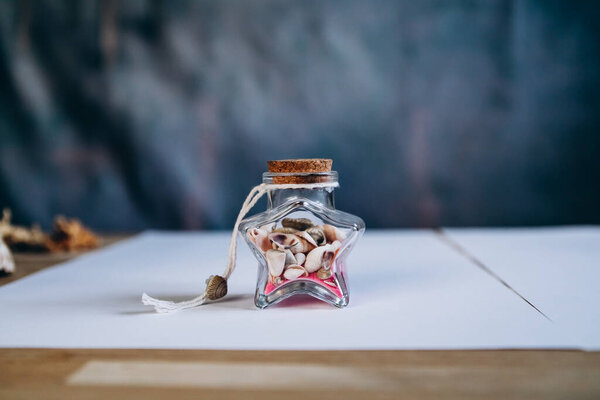 A glass jar in the shape of a star inside pink sand and small shells on a blue background close-up. Souvenir. Maritime concept.