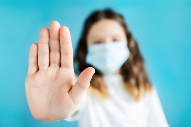 Mavi tıbbi maskeli genç bir kız, beyaz tişörtlü, elini öne uzatıyor ve mavi arka planda dur işareti yapıyor. Koronavirüs pandemisi sırasında antiviral koruma.