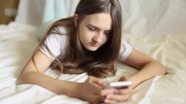 Mutlu, rahat, akıllı bir kız elinde cep telefonuyla geziyor, komik bir video izliyor, gülüyor, yatakta uzanıyor, gülümsüyor, sosyal ağlarda mutlu bir şekilde sohbet ediyor, evdeki yatakta dinleniyor.