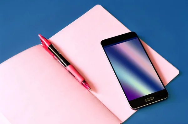 Smartphone with abstract holographic flow on display and pink notebook on classic blue background — 스톡 사진