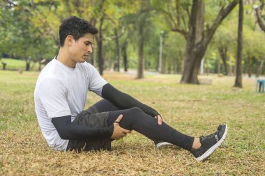Parkta koşan genç erkek sporcular dizlerinde ağrı hisseder ve oturur. Elleri dizinde tutarlar..