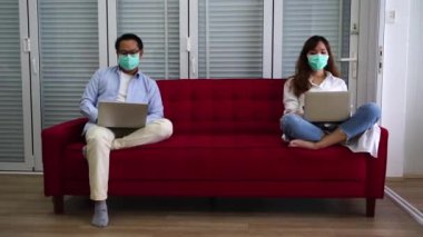 Maske takan koca ve maske takan Asyalı eş dizüstü bilgisayarla çalışıyor kırmızı koltukta oturuyor evde iş görüşmeleri yapıyor. Sosyal mesafe ve ev kavramından çalışma..