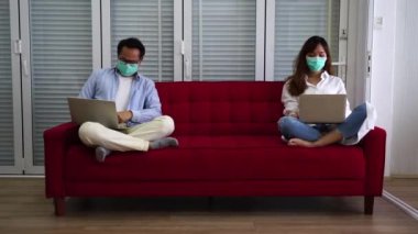 Maske takan koca ve maske takan Asyalı eş dizüstü bilgisayarla çalışıyor kırmızı koltukta oturuyor evde iş görüşmeleri yapıyor. Sosyal mesafe ve ev kavramından çalışma..