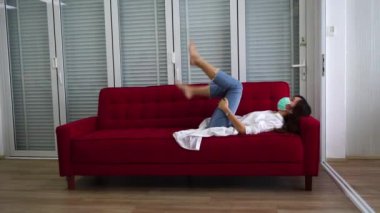 Asyalı kadınlar kot pantolon giyer, maske takar, kanepede bacak kaldırır, evin oturma odasında egzersiz yapar. Ev konseptinde karantina ve egzersiz yapar..