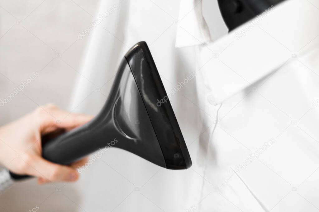 the girl holds in her hand a steamer for clothes for white shirts that hang on a hanger