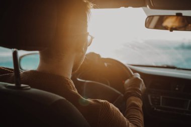 Otoyolda araba süren bir adam var. Batan güneş ön camda parlıyor.