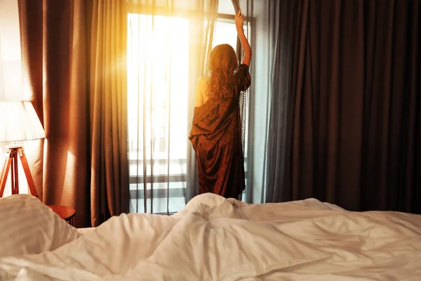 A woman meets the dawn from the window of her bedroom, opening the curtains in a silk long dressing gown