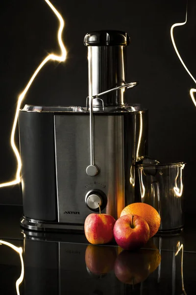 Freshly squeezed juice from a juicer ready for use from young carrots and red apples and juicy oranges standing on a black mirror image background
