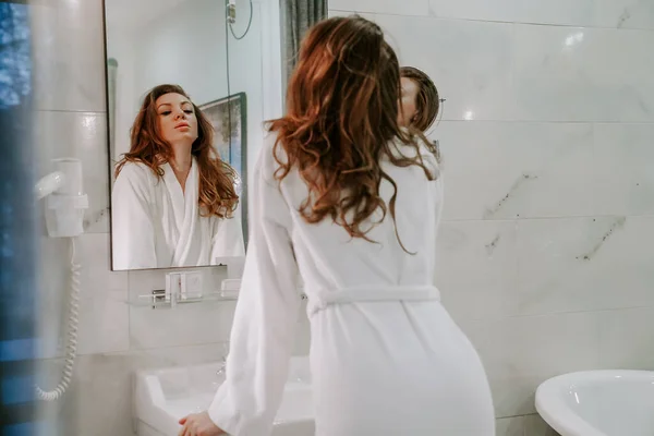 Duştan sonra beyaz bornozlu güzel bir kadın banyodaki aynaya bakar. Ev yapımı vücut bakımı. spa tedavileri