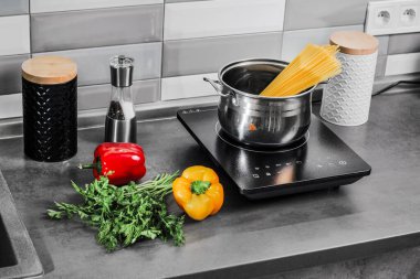 Endüksiyon sobasında tavada pişmiş spagetti. Mutfak masasında maydanozlu kırmızı ve yeşil biber. Kadın yemek yapıyor.