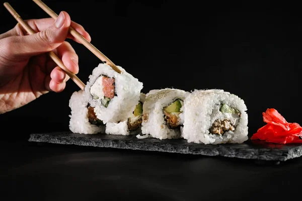 a man eats chopsticks hosomaki rolls with salmon and butter cream and black sesame seeds on a black background. japanese food close-up. veggie dish