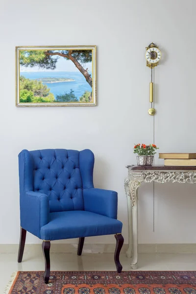 Interior composition of retro blue armchair, vintage wooden beige table, and pendulum clock over off white wall, tiled beige floor and orange ornate carpet