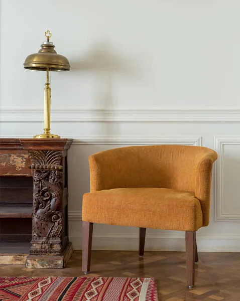 Sillón naranja y aparador adornado vintage de madera y lámpara de mesa en la pared de color blanco hueso y alfombra adornada sobre suelo de parquet de madera — Foto de Stock