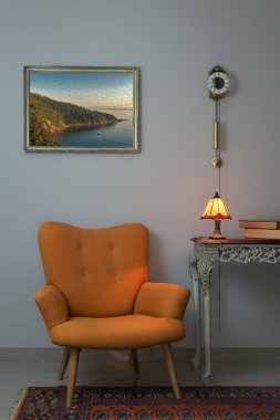 Retro turuncu tekli, vintage ahşap bej masa, ışıklı antika masa lambası, eski kitaplar ve sarkaç saatin üzerine beyaz duvar ve turuncu halı iç atış