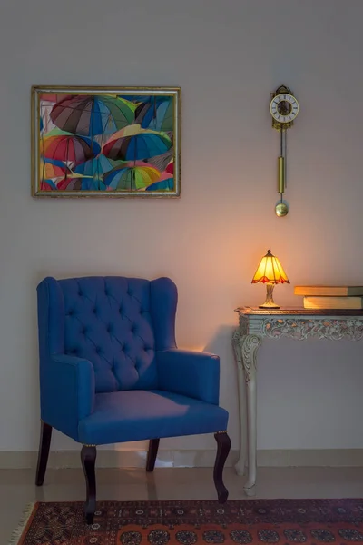 Composizione interna di poltrona blu, tavolo beige vintage in legno, lampada da tavolo illuminata, libri, orologio a pendolo e tappeto decorato arancione — Foto Stock