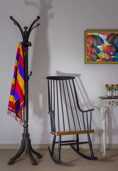 Внутренняя съемка винтажного кресла-качалки, вешалки для пальто и цветочного горшка на старинном старинном столе на фоне белой стены с одной повешенной картиной — стоковое фото