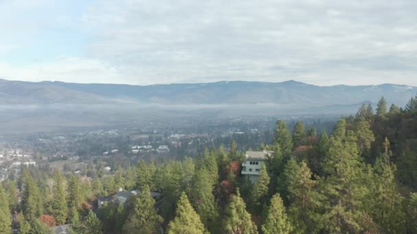 Volviendo sobre árboles y casas en la colina con la ciudad y las montañas como fondo — Vídeos de Stock