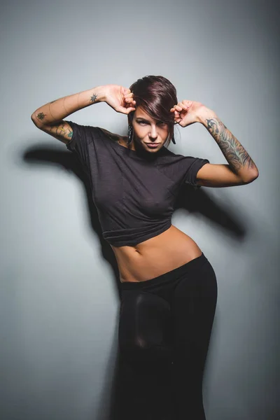 Bella donna bruna con tatuaggi e una figura sportiva in posa su sfondo scuro in Studio — Foto Stock