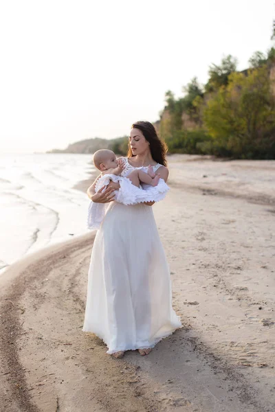 Uma jovem mãe segurando um bebê em seus braços na praia junto ao mar — Fotografia de Stock