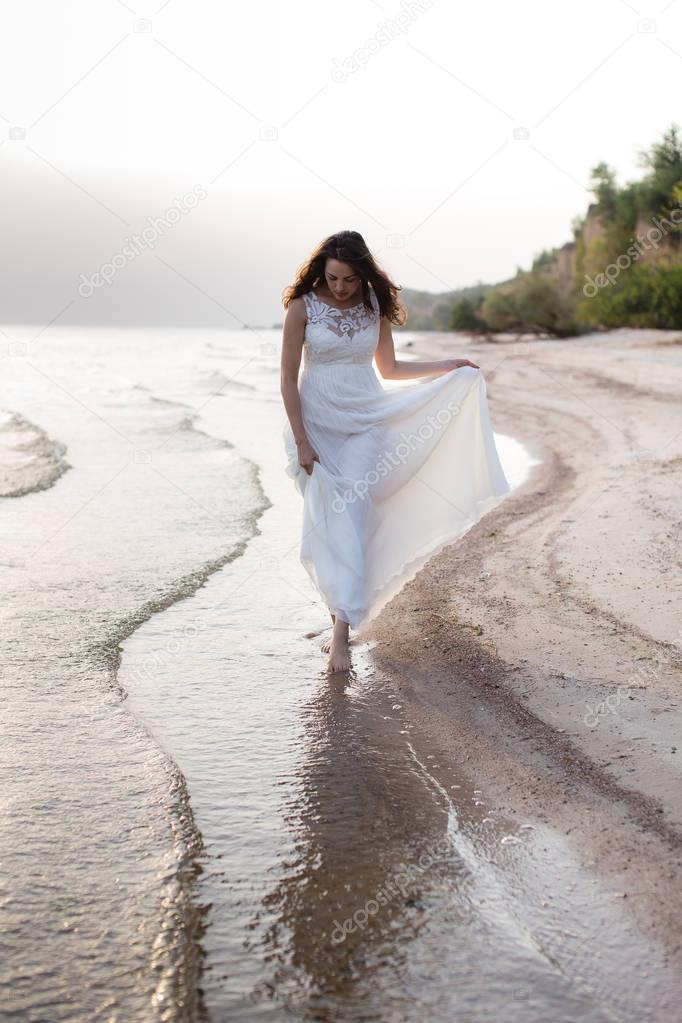 Beautiful young brunette woman in white long dress at the sea shore