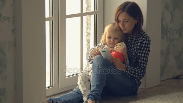 Madre con asientos de hija pequeña cerca de la ventana y utiliza el teléfono inteligente — Vídeo de stock