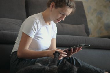 Güzel beyaz kadın evde yerde oturmuş dijital tablet bilgisayar kullanıyor ve siyah kedisine sataşıyor.