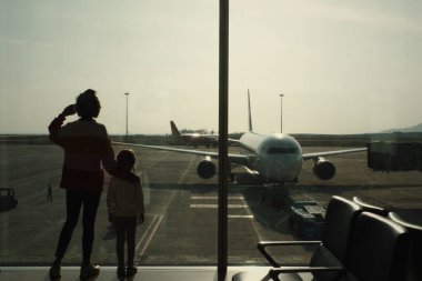 Annesinin ve kızının siluetleri, havaalanındaki bekleme odasında, terminal penceresinde duruyor. Annem kahveyi kağıt bardakta içer.